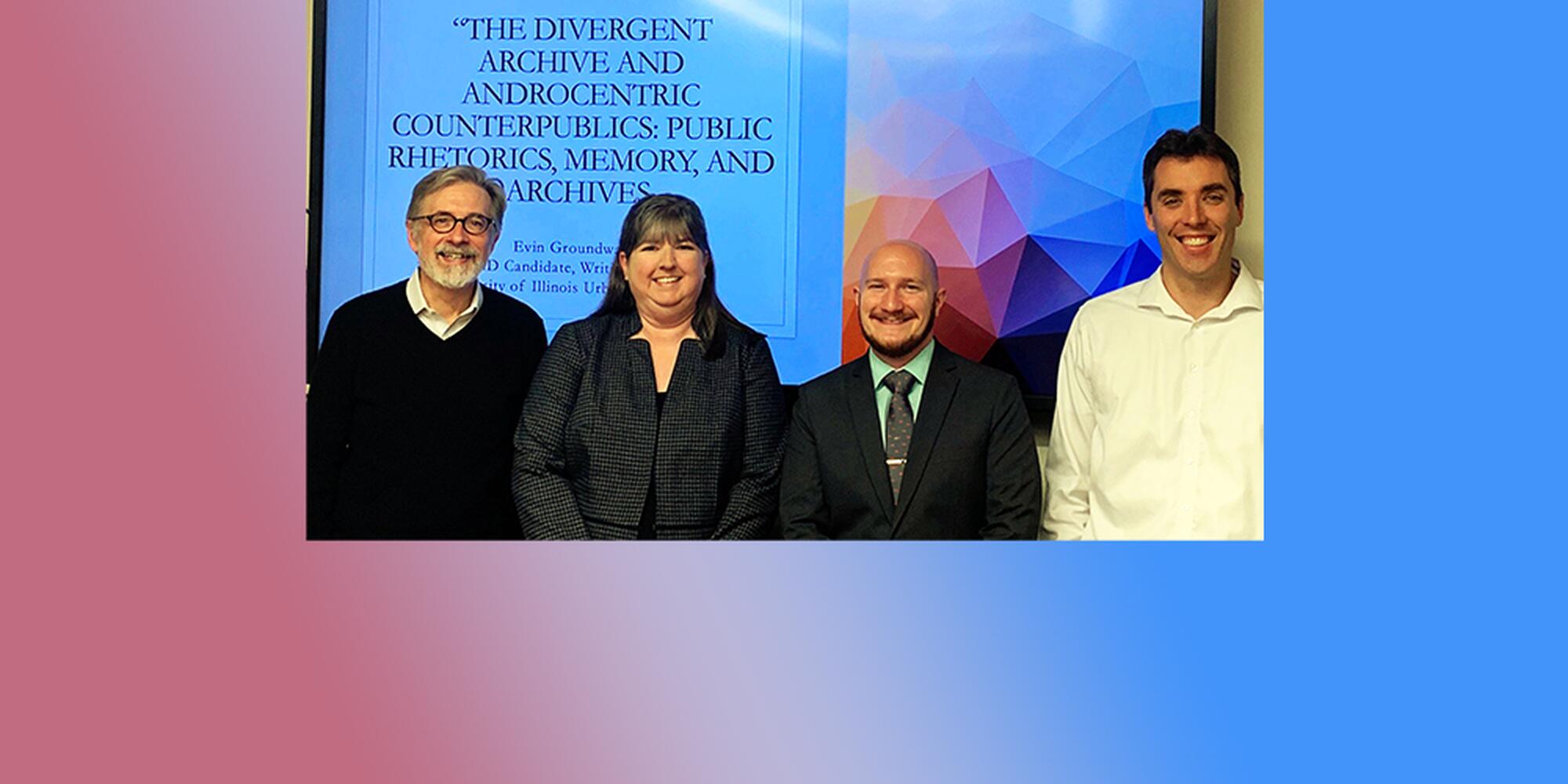 Four white people stand side by side against a cover slide for a dissertation defense; the slide reads "The Divergent Archive and Androcentric Counterpublics: Public Rhetorics, Memory, and Archives." One of the people present is a PhD candidate who just defended his work; the other three are professors (a man with grey hair, a beard, sweater, and glasses; a woman wearing a blouse; and a man wearing a white dress shirt).